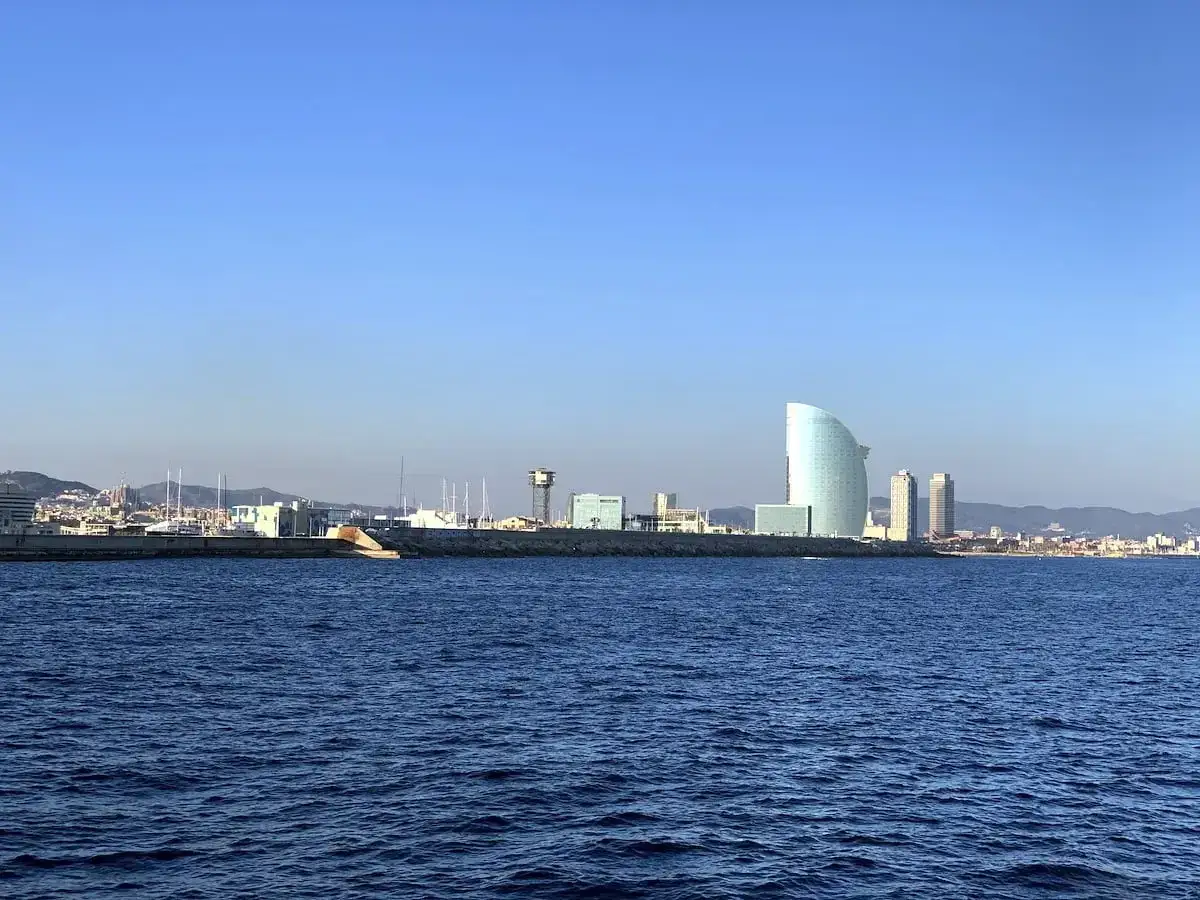 Widok na nowoczesną panoramę miasta z charakterystycznym budynkiem w kształcie żagla. To właśnie tutaj jest Barcelona, z błękitnym morzem na pierwszym planie.