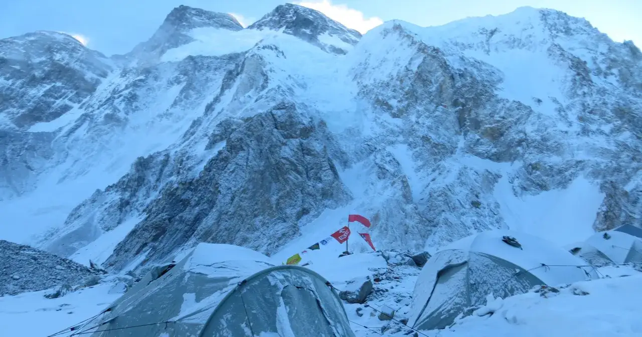 Namioty w śniegu u podnóża majestatycznego, ośnieżonego broad peak szczytu. W tle powiewają flagi.