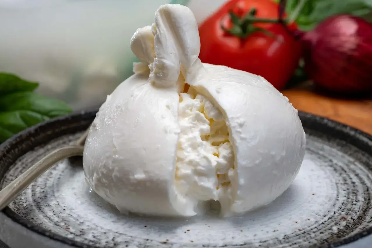 Una burrata fresca, con il suo cuore cremoso che fuoriesce, pronta per essere gustata. Sullo sfondo, pomodoro, cipolla rossa e basilico.