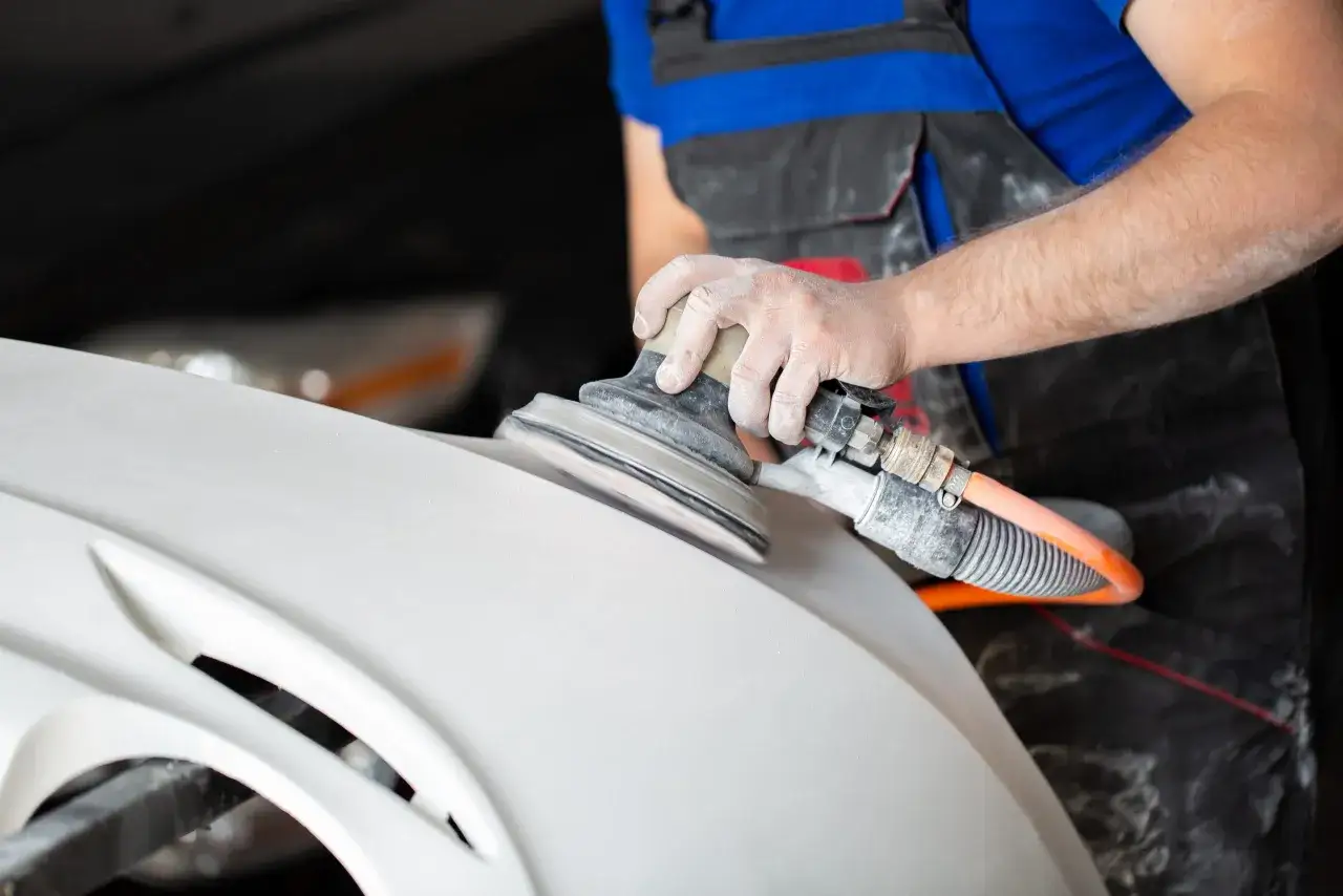 Main d'un ouvrier utilisant une ponceuse pour le d&eacute;capage m&eacute;canique d'une pi&egrave;ce automobile blanche.