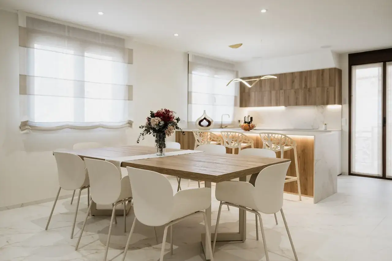 Elegantes cocinas abiertas al sal&oacute;n con barra, mesa de comedor y sillas blancas, creando un espacio moderno y luminoso.