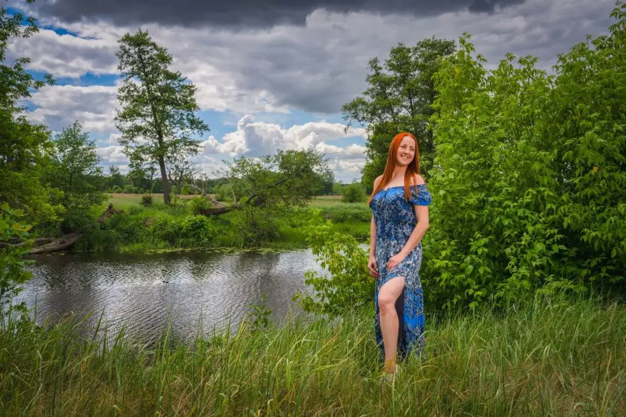 Ruda kobieta w niebieskiej sukience stoi w trawie nad rzeką. W plenerze, wśród zieleni i chmur, czuje się wolna.