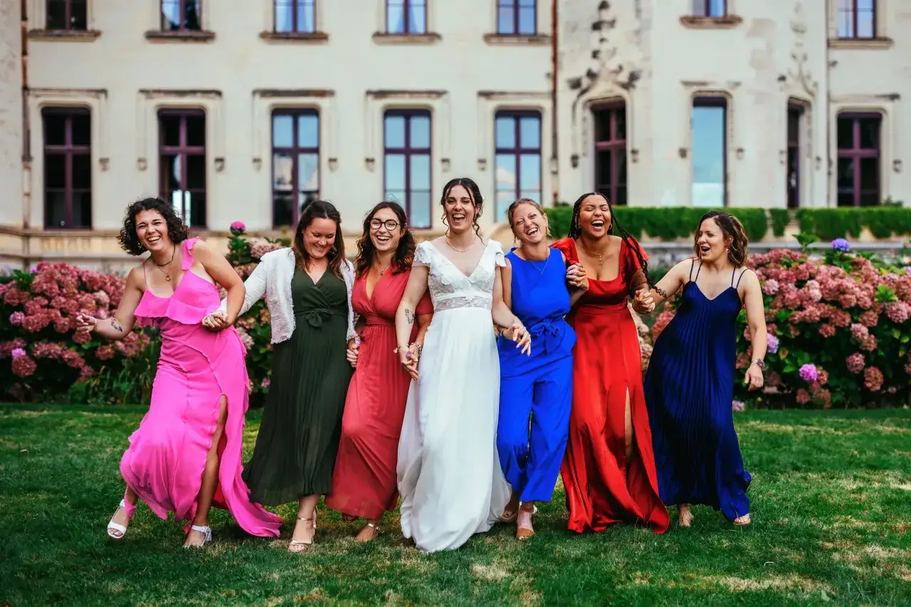 Un groupe de femmes rit joyeusement devant un château. La mariée en blanc est entourée de ses demoiselles d'honneur, chacune dans une robe colorée. La différence entre témoin et demoiselle d'honneur est subtile, mais leur complicité est évidente.