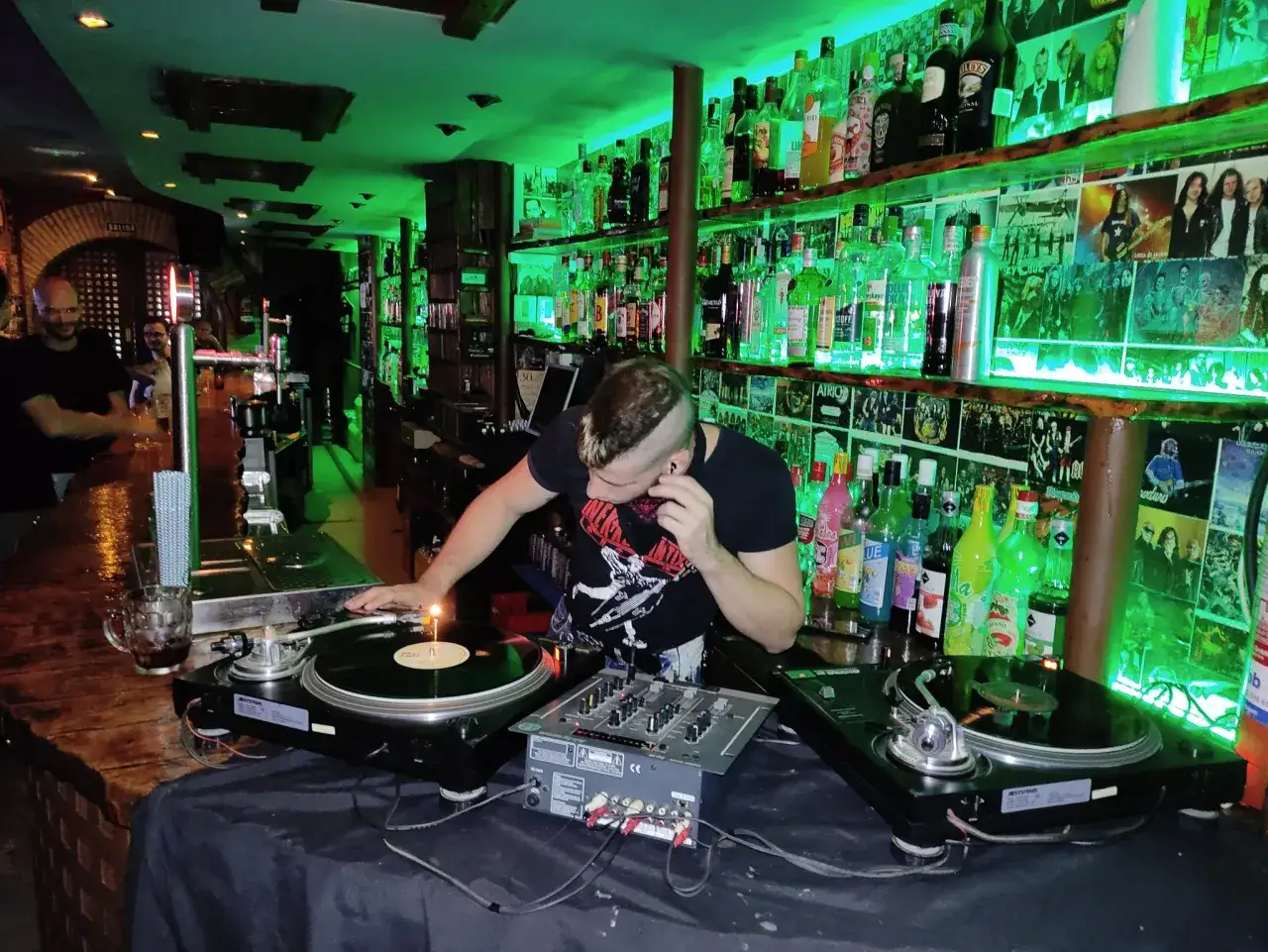 DJ en linacero cafe zaragoza, con luces verdes y botellas de licor en estanterías.