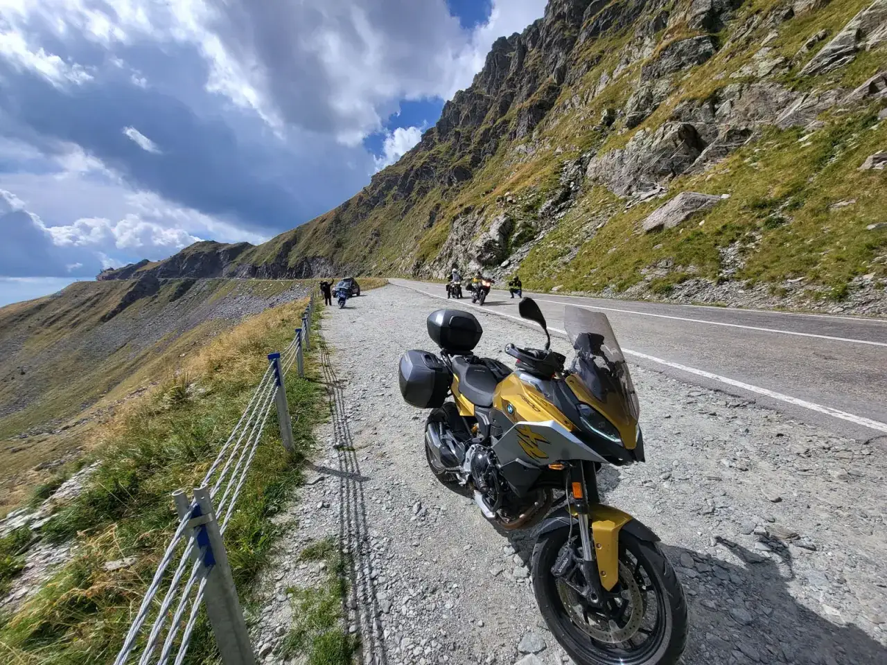 Złoty motocykl na żwirowym poboczu malowniczych dróg Rumunii, w tle góry i chmury.