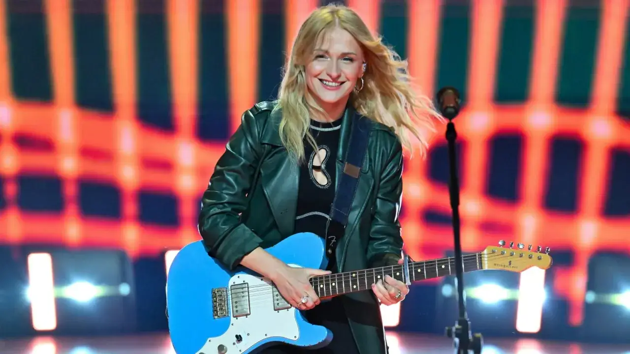 Uśmiechnięta polska piosenkarka z blond włosami gra na niebieskiej gitarze elektrycznej na tle pomarańczowych świateł.
