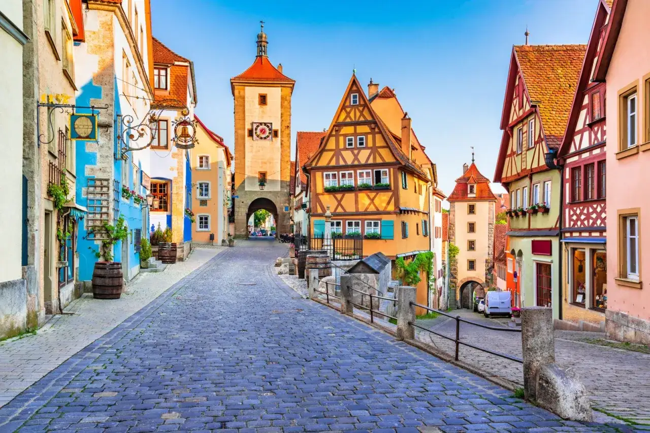 Bunte Fachwerkh&auml;user s&auml;umen eine Kopfsteinpflasterstra&szlig;e, die zu einem mittelalterlichen Stadttor f&uuml;hrt. Ein malerischer Anblick historischer Orte in Deutschland.
