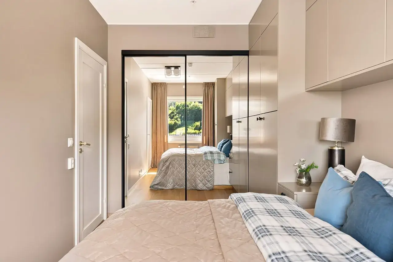 Ein großer spiegel im schlafzimmer reflektiert das Bett, die Gardinen und das Fenster mit Blick ins Grüne.