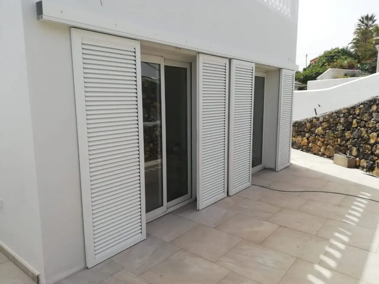 Una fachada blanca con una contraventana corredera de lamas blancas, que protege un ventanal de cristal.