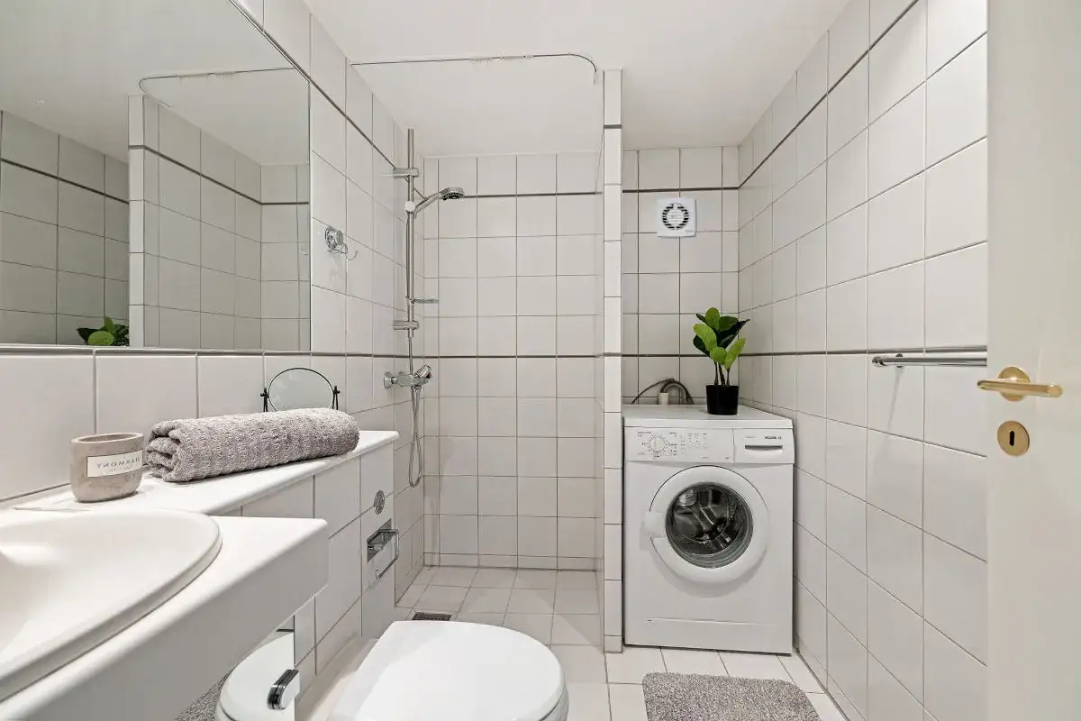 Ba&ntilde;o con lavadora, ducha y espejo. Se ve un extractor de ba&ntilde;o en la pared, ideal para mantener el ambiente fresco.