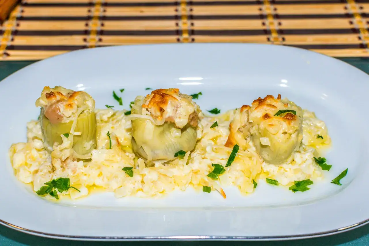Tres alcachofas gratinadas con queso cremoso y perejil picado, servidas en un plato blanco.
