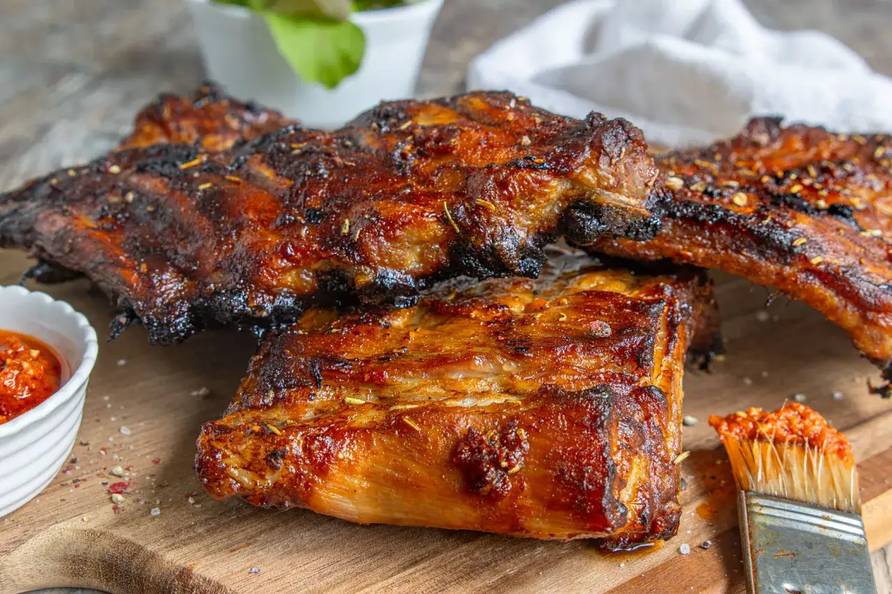 Saftige Spareribs, perfekt gegrillt, liegen auf einem Holzbrett. Eine kleine Schale mit scharfer Sauce und ein Pinsel mit Sauce daneben deuten auf die Zubereitung hin. Die Frage 