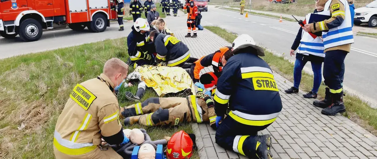 Pierwszy ratownik z PSP udziela pomocy poszkodowanemu na miejscu zdarzenia. W tle wóz strażacki i inni ratownicy.