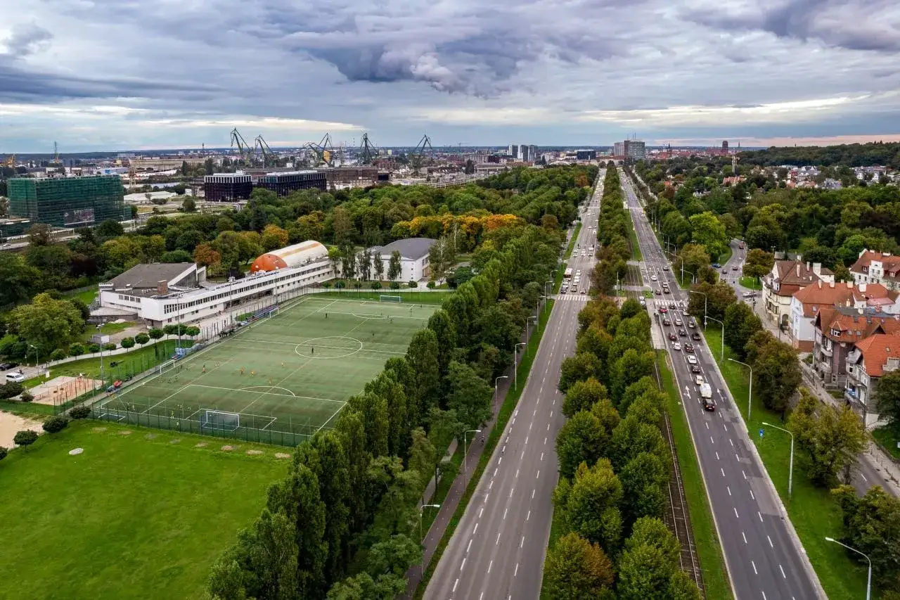Widok na boisko i ulicę w Gdańsku Aniołkach. W tle portowe dźwigi i zabudowa miejska.