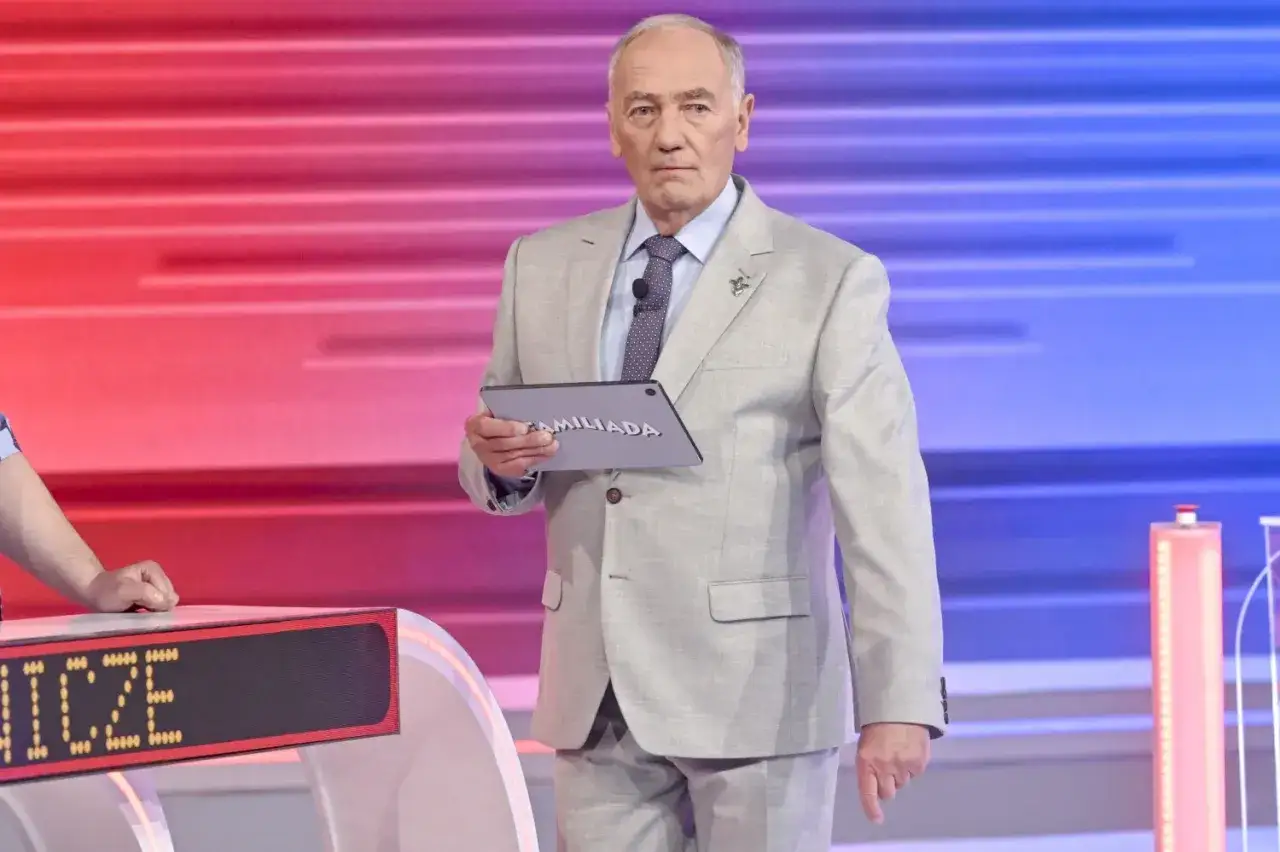 Prowadzący Familiady, ubrany w jasny garnitur, trzyma tablet z napisem