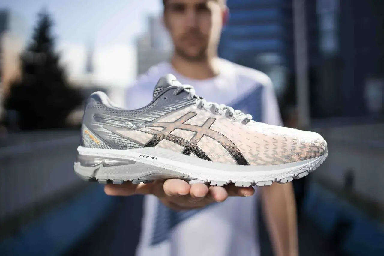 En person håller upp en ljusrosa och grå löparsko, Asics Gel Luminus 2 dam, med stadsmiljö i bakgrunden.