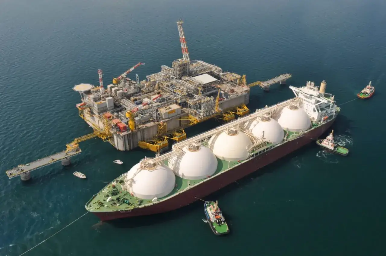 Praca na statku LNG: potężna jednostka z kopułami ładunkowymi przy platformie wydobywczej, holowniki asystują.