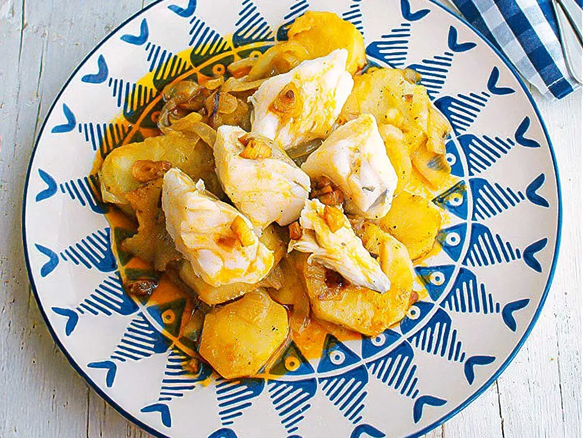 Colas de rape al horno con patatas y cebolla, servido en un plato decorado con peces azules.