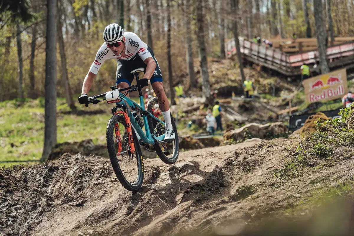 Kolarz w akcji podczas zawod&oacute;w MTB, pokonujący błotnisty zjazd. W tle widać kibic&oacute;w i logo Red Bull.