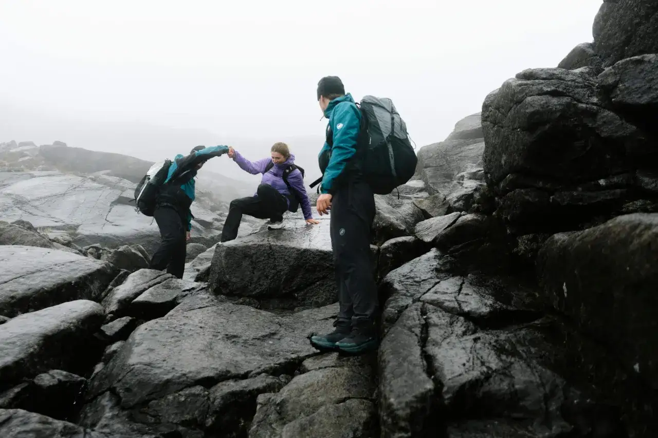 Drei Wanderer k&auml;mpfen sich durch nasses Gestein. Ein Mann reicht einer Frau die Hand, w&auml;hrend ein weiterer Mann im Vordergrund steht. Ein Gewitter in den Bergen zieht auf.