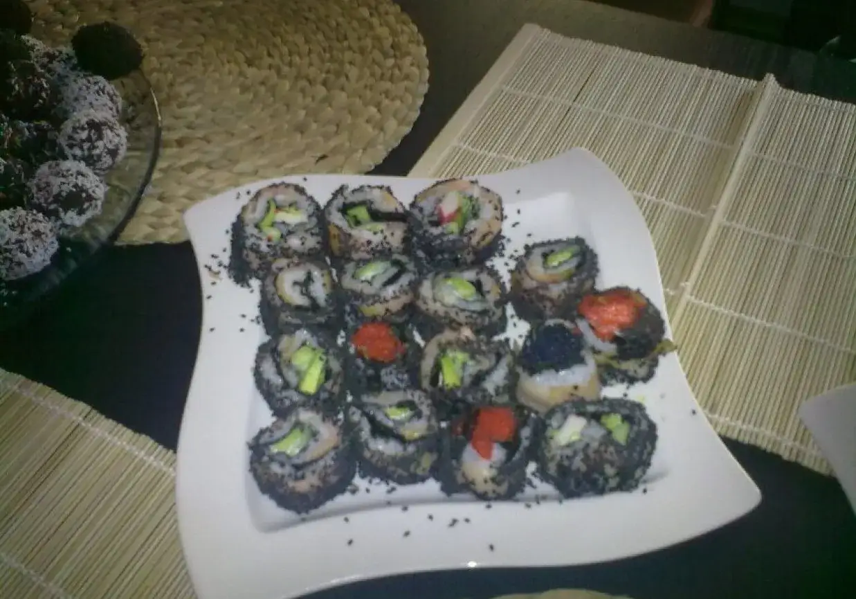 Talerz pełen sushi z sezamem, z widocznymi kawałkami awokado i ikrą. Obok deserki w wiórkach kokosowych.