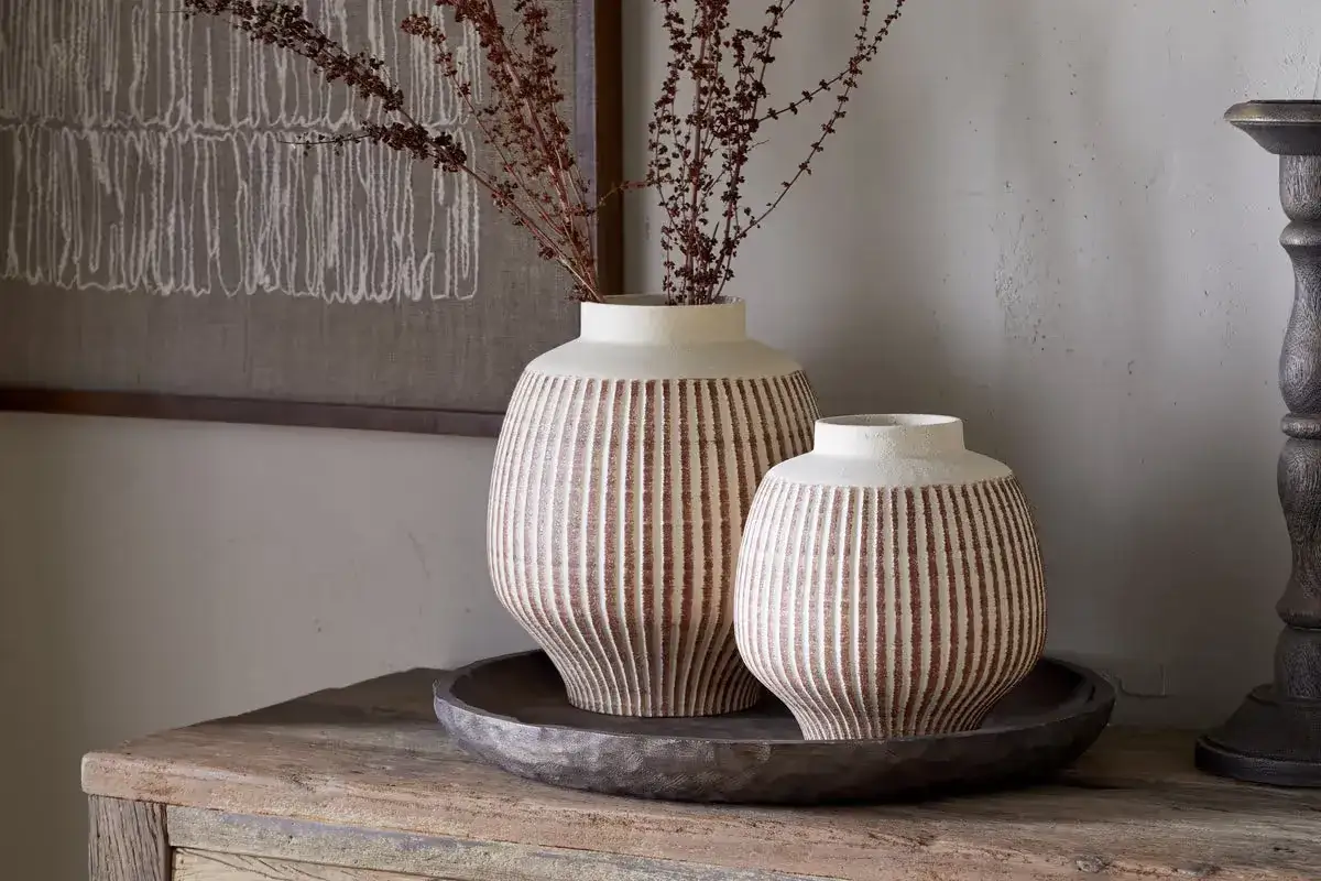 Zwei gestreifte Vasen, eine größer als die andere, sind auf einer Holzplatte arrangiert. Sie zeigen, wie man eine Vase dekorieren kann, ohne Blumen zu verwenden, mit getrockneten Zweigen.