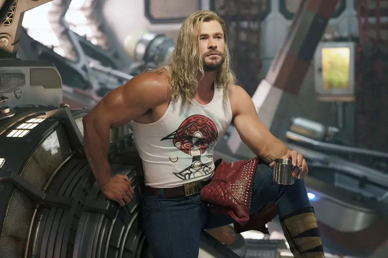 Thor w białej koszulce z nadrukiem, jeansach i czerwonych butach, siedzi na tle futurystycznego wnętrza, trzymając kubek.
