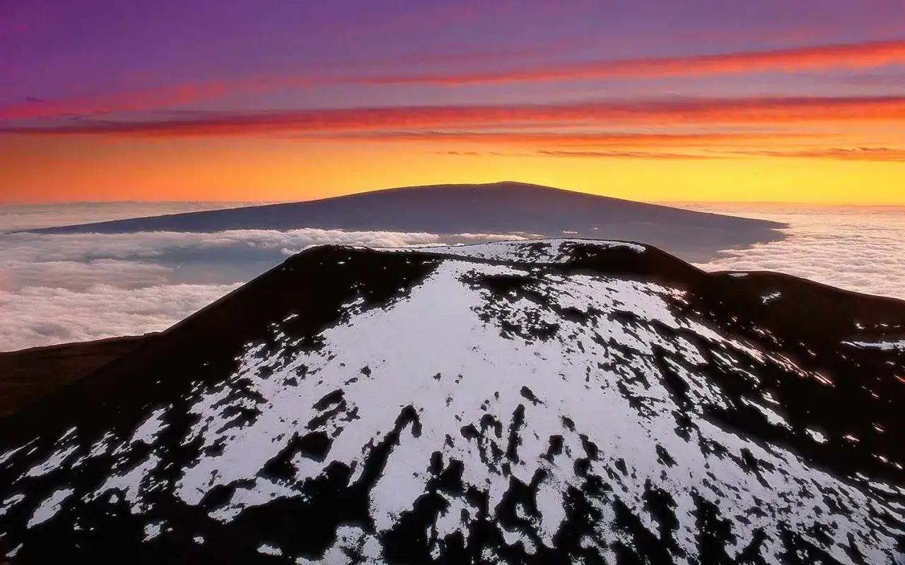 Szczyt Mauna Kea, największego wulkanu na świecie, pokryty śniegiem, na tle purpurowo-pomarańczowego nieba i morza chmur.