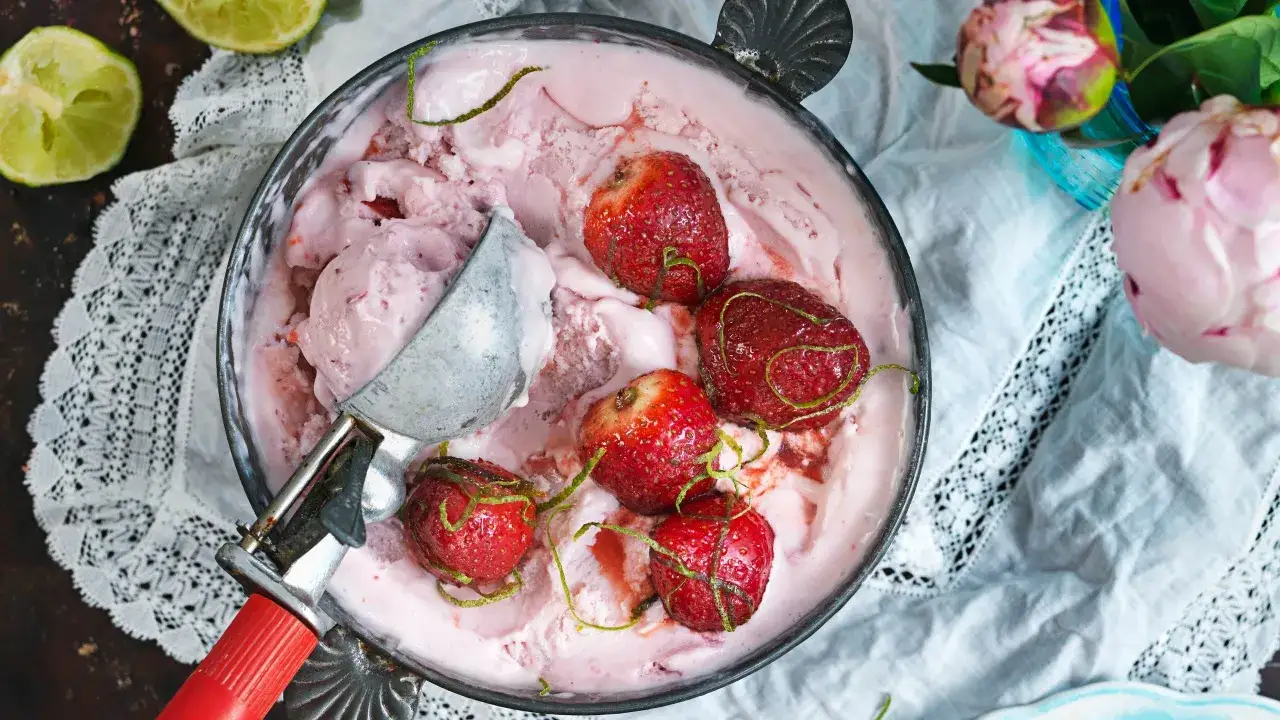 En skål med rosa jordgubbsglass, toppad med hela jordgubbar och rivet limeskal. En glasskopa vilar i skålen.