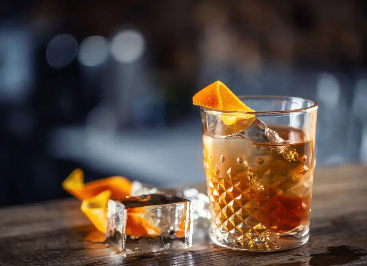 Gotowy drink Old Fashioned w szklance typu rocks z dużą kostką lodu i skórką pomarańczy