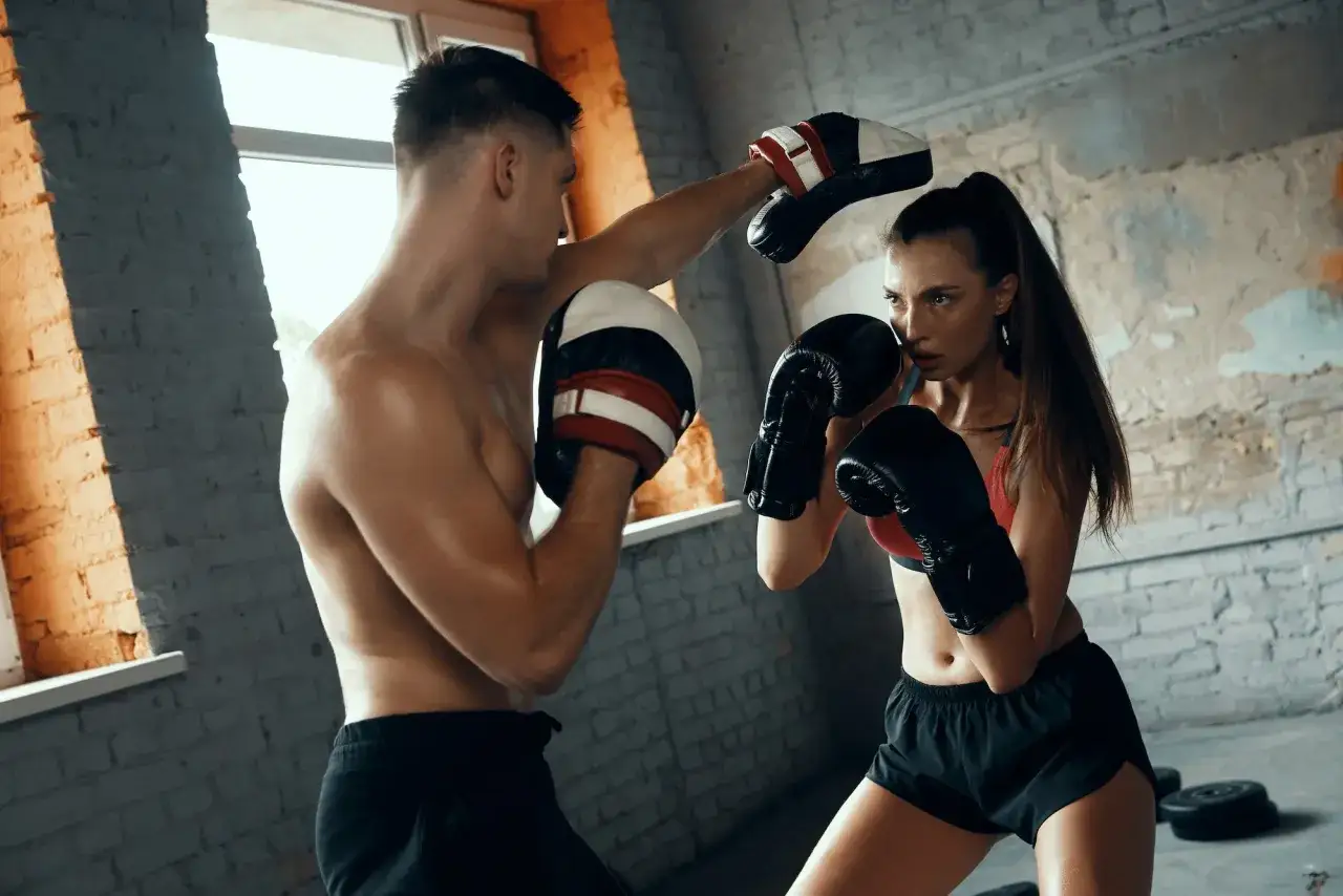 A man and woman train for a boxing match in a gritty gym. He holds pads as she throws a punch, both focused and determined.