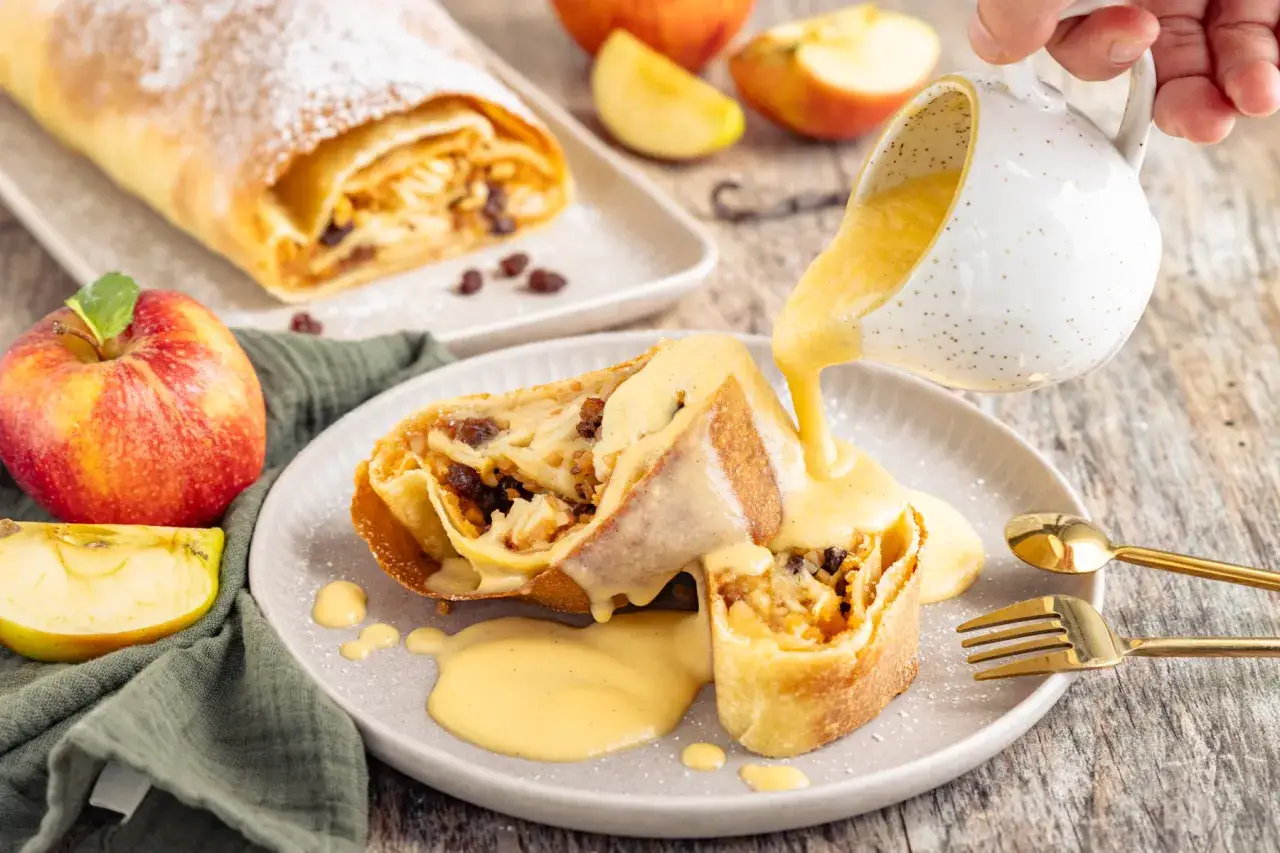 Ein Stück Apfelstrudel wird mit cremiger Vanillesauce aus Puddingpulver übergossen. Daneben liegen Äpfel und Rosinen.