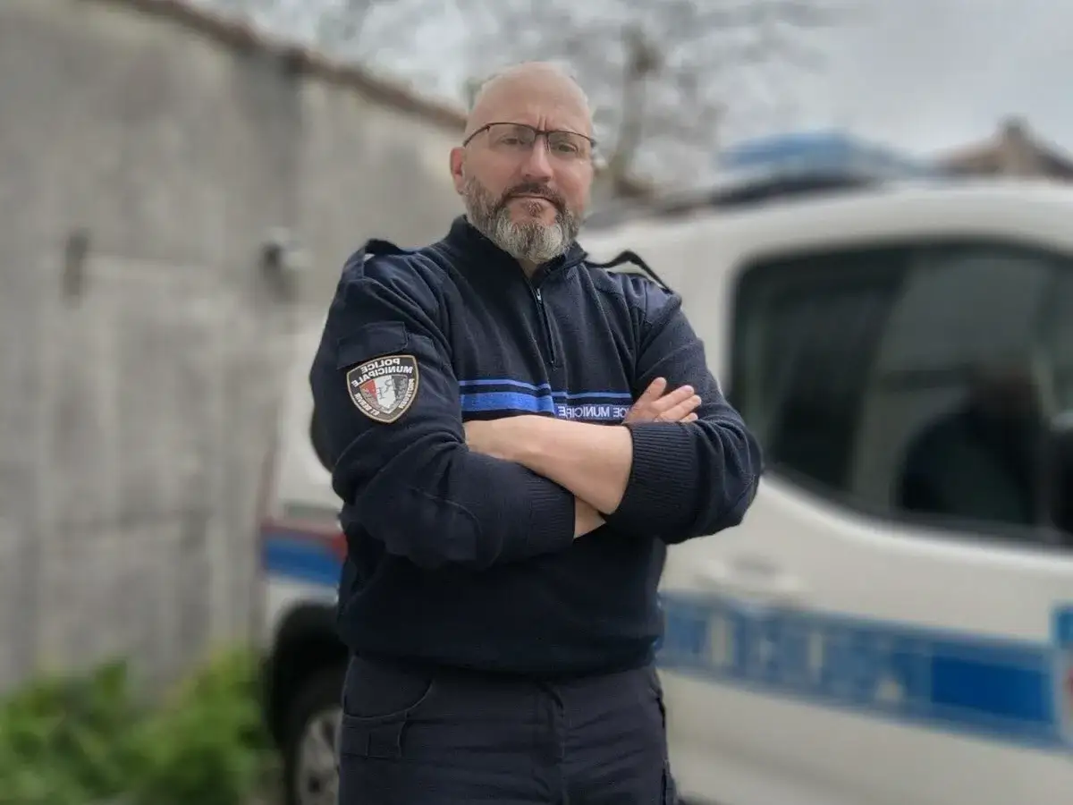 Policier municipal, bras croisés, prêt pour une lettre de motivation police municipale mutation. Véhicule de service flouté en arrière-plan.