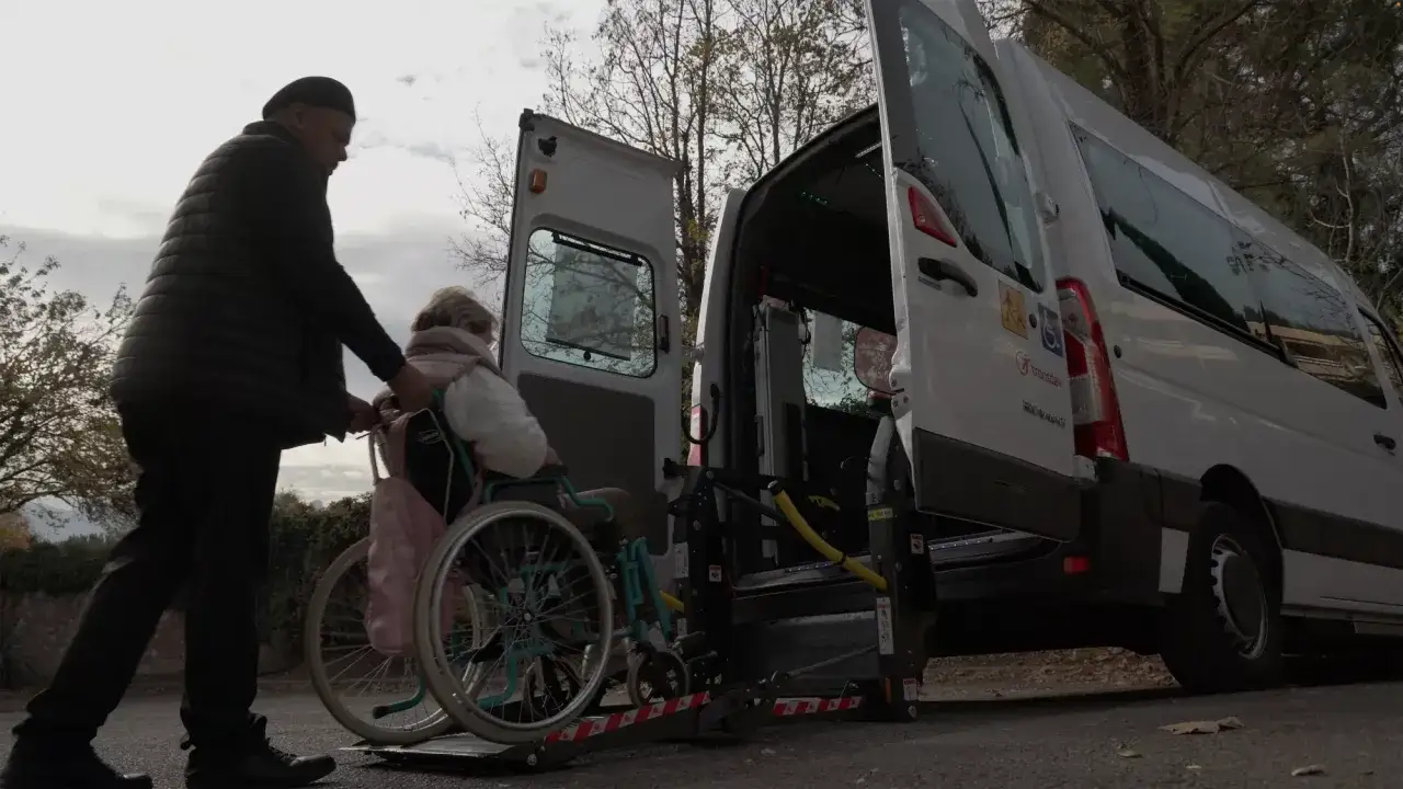 Un uomo spinge una donna in sedia a rotelle verso un minibus con pedana. Servizio di trasporto accessibile, 104 condivisa.