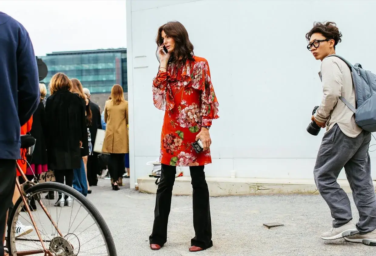 Donna con vestito floreale rosso e pantaloni neri sotto, parla al telefono. A destra, un fotografo con zaino.