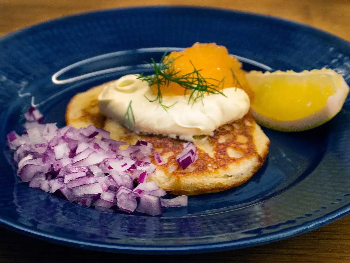 Blinier recept: Små, gyllene pannkakor toppade med gräddfil, rom, dill och en citronklyfta. Serveras med finhackad rödlök.