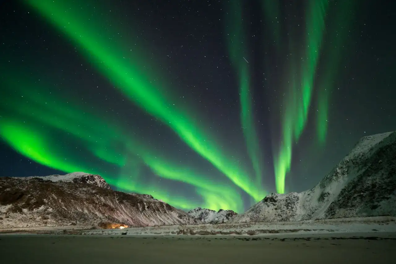 Grönt norrsken dansar över snöklädda berg vid en strand på Lofoten.
