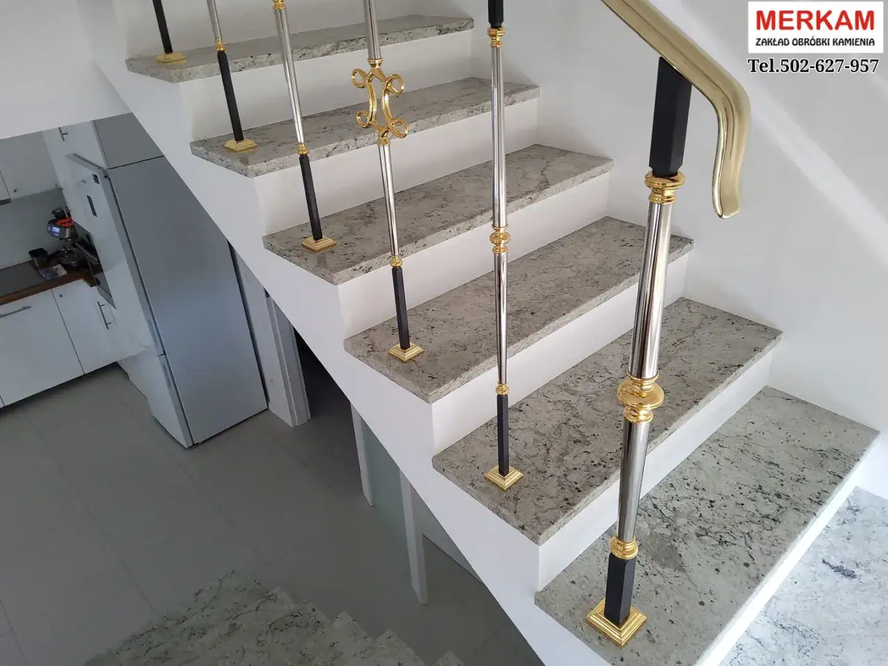 Schody z kamienia aglomerat z elegancką, złotą balustradą. W tle kuchnia z lodówką.