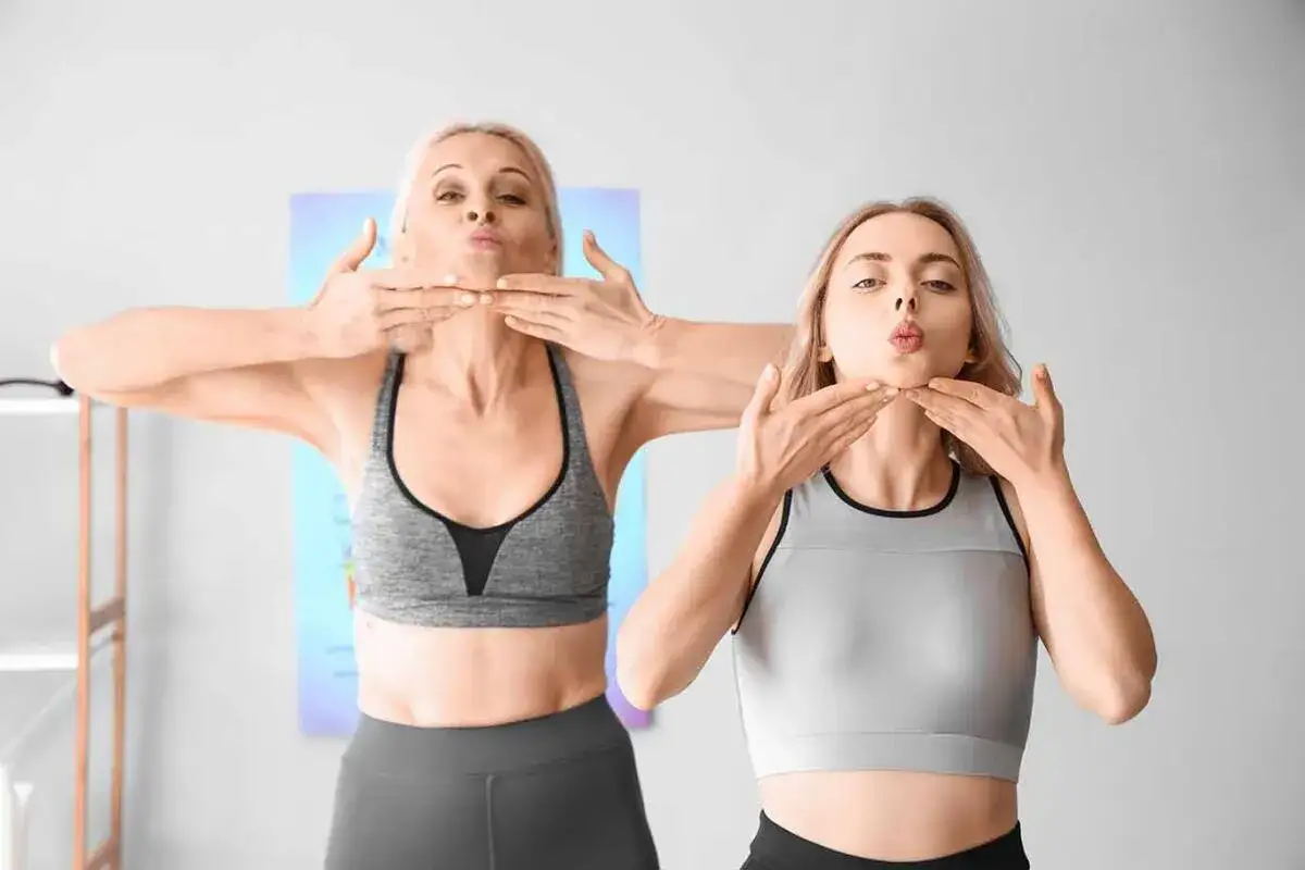 Two women practice face yoga, puckering their lips. While face yoga is often promoted for anti-aging, some potential side effects include temporary muscle soreness or bruising.