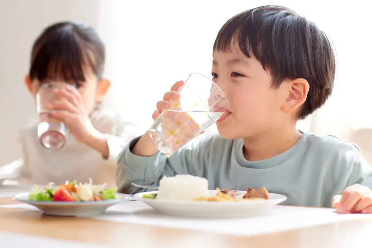 Zwei Kinder essen und trinken Wasser. Der Junge im Vordergrund trinkt aus einem Glas, während das Mädchen im Hintergrund ebenfalls ein Glas hält.