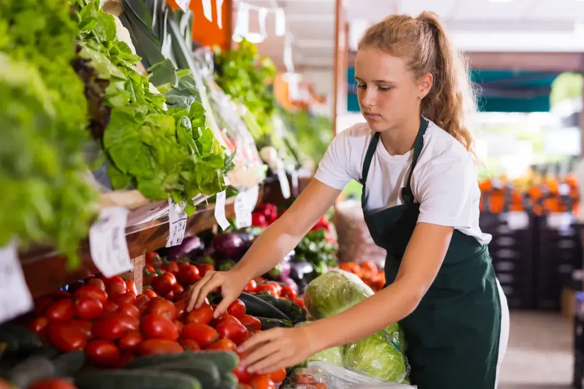 Młoda dziewczyna w fartuchu układa pomidory na stoisku warzywnym. To jej praca dla nieletnich, gdzie uczy się obsługi klienta i dbania o świeżość produktów.