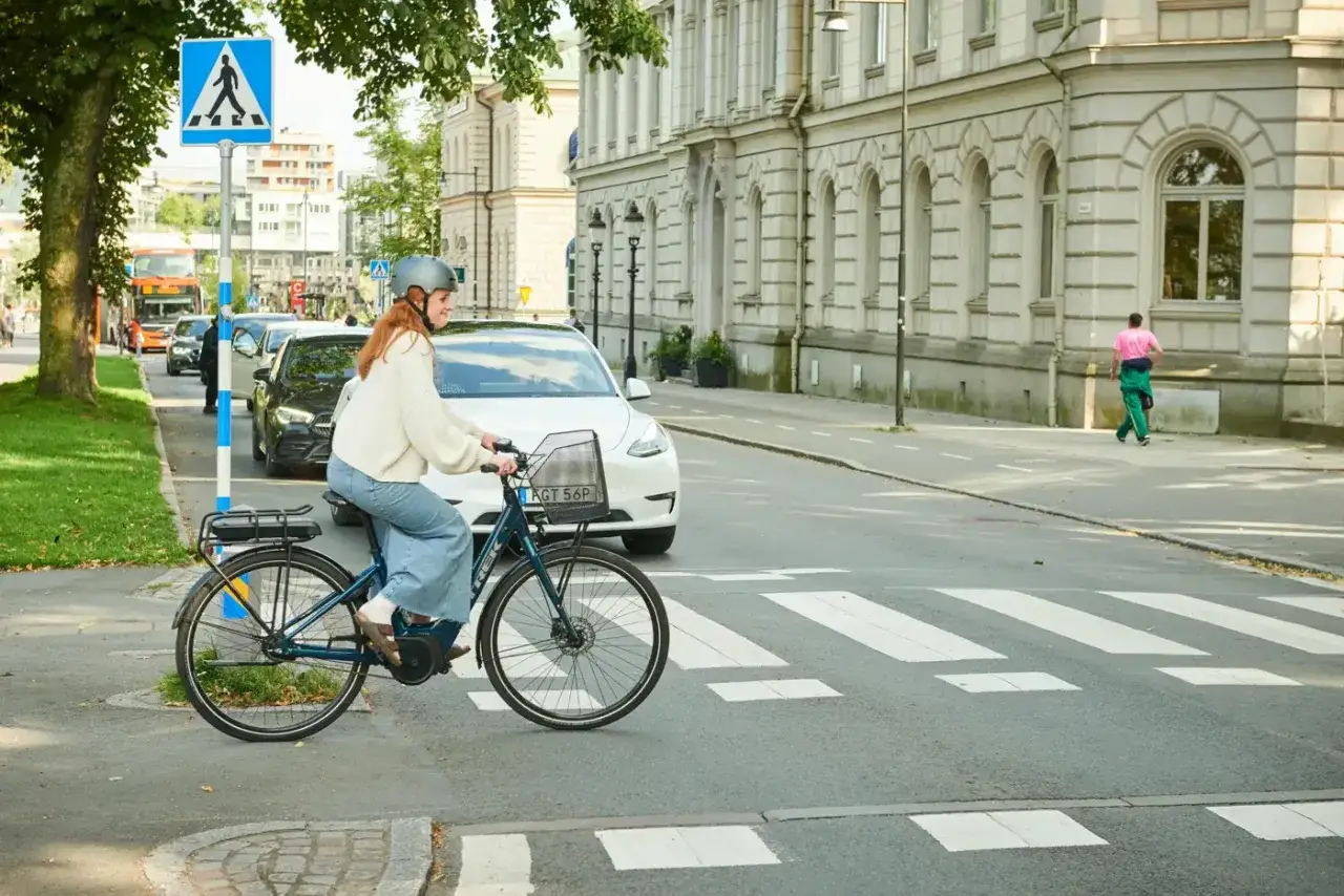 En kvinna p&aring; elcykel korsar ett &ouml;verg&aring;ngsst&auml;lle. Hon b&auml;r hj&auml;lm. Trafik och byggnader syns i bakgrunden. Elcykel regler &auml;r viktiga f&ouml;r s&auml;kerheten.