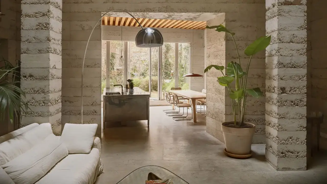 Interior de una casa entre medianeras con paredes de hormig&oacute;n texturizado, sof&aacute; blanco, cocina y comedor con vistas al jard&iacute;n.