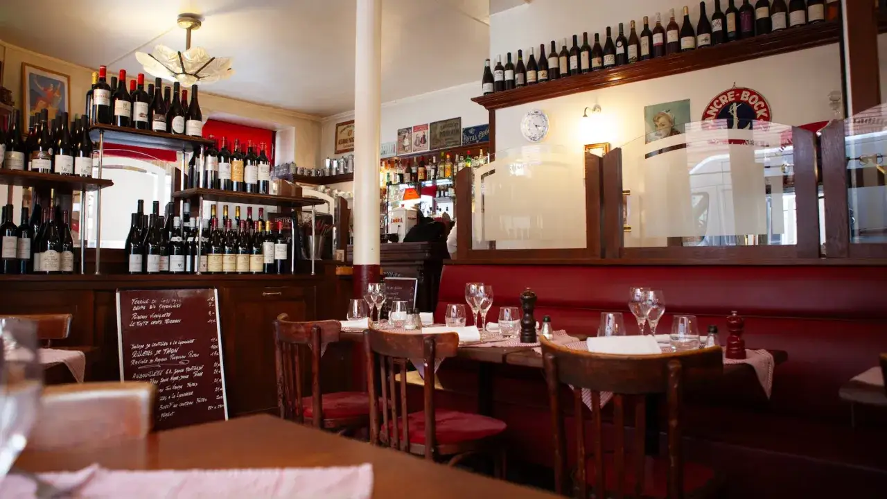 Intérieur d'un bistrot parisien avec des étagères remplies de bouteilles de vin, tables dressées, banquettes rouges. L'ambiance évoque le plus vieux restaurant de Paris.