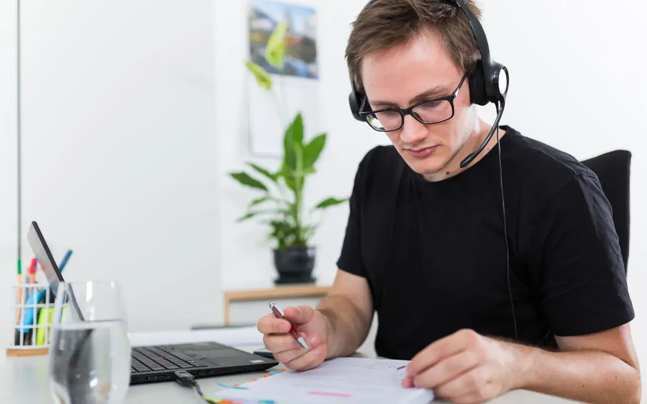 Mann mit Headset lernt für seinen **bachelor berufsbegleitend**. Er sitzt am Schreibtisch, vor ihm ein Laptop und Notizen.