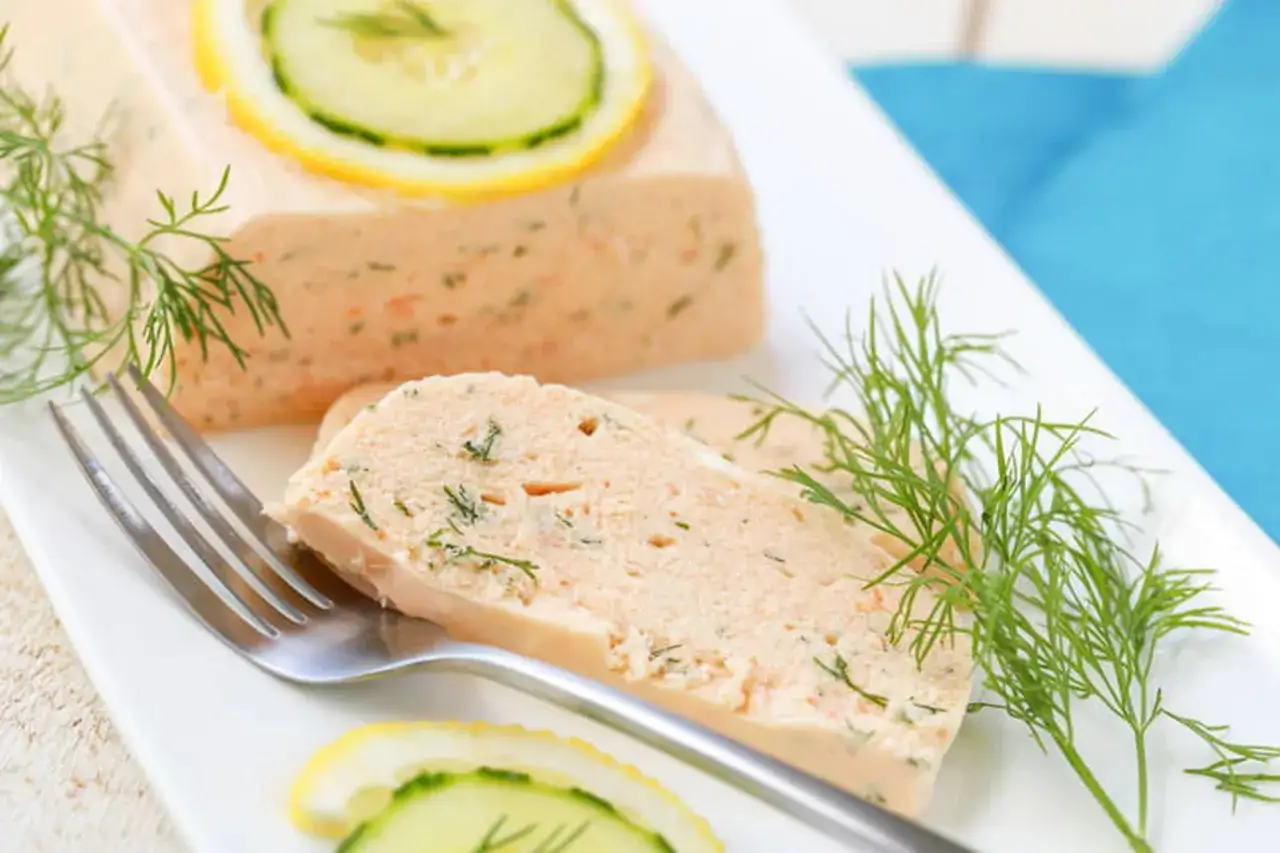 Une tranche de terrine de saumon, garnie d'aneth et de rondelles de concombre et de citron, attend sur une assiette blanche avec une fourchette.