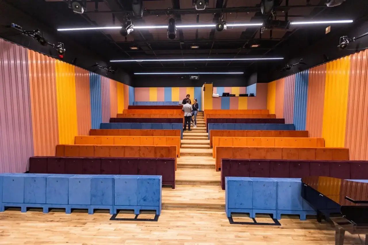 Kolorowe siedziska i ściany tworzą nowoczesną przestrzeń, idealną dla **teatru ludowego w Krakowie**. Trzy osoby rozmawiają na schodach.