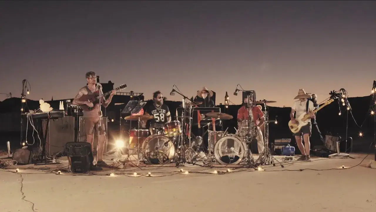 Un groupe joue de la musique en plein air au coucher du soleil. Les membres du groupe, **thee oh sees**, sont entour&eacute;s de lumi&egrave;res d&eacute;coratives.