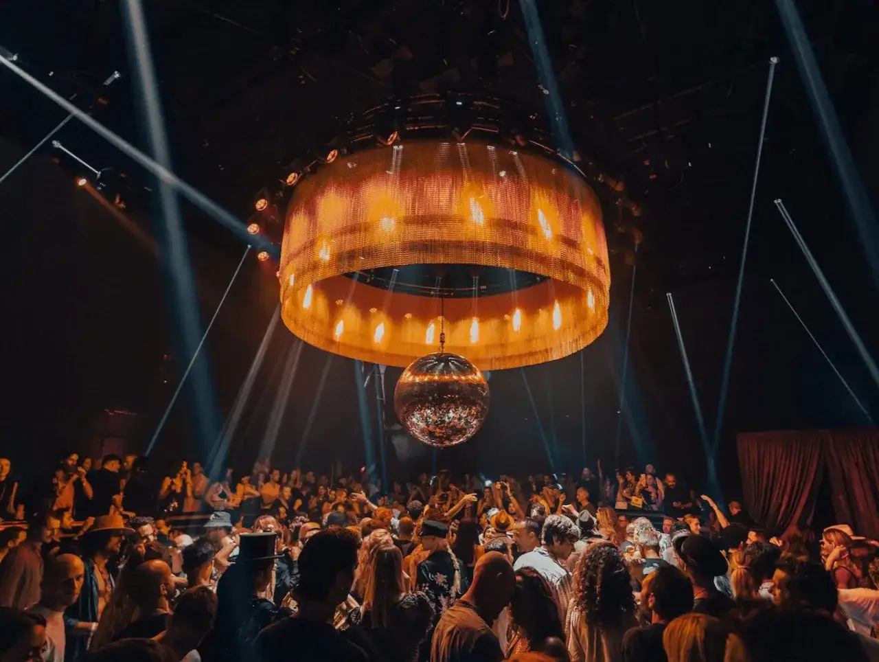 Folla in delirio sotto una sfera stroboscopica e un'imponente lampada dorata. L'atmosfera &egrave; elettrica, perfetta per l'apertura discoteche Ibiza.