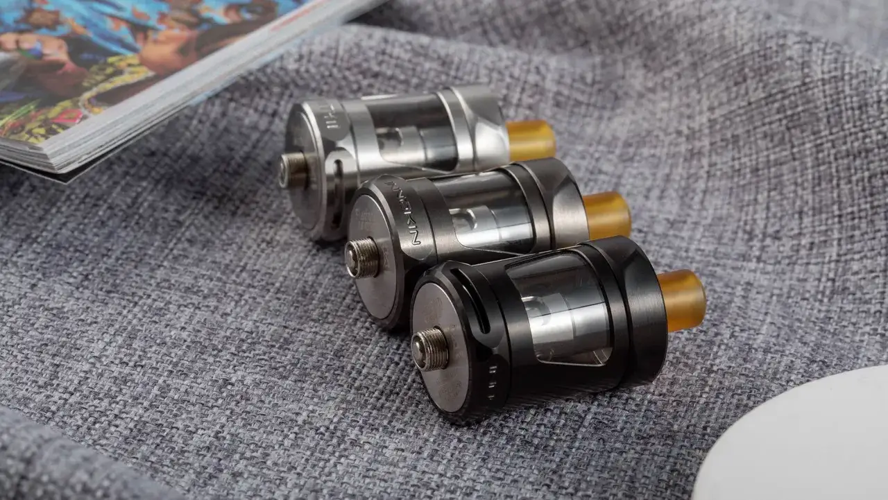Tres atomizadores GTX Tank 22 en plata, gris y negro, junto a una revista sobre tela gris.